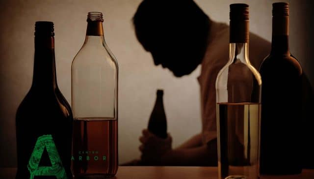 Hombre cabizbajo sosteniendo una botella, rodeado de otras botellas de alcohol, con el rostro desenfocado y en penumbra.