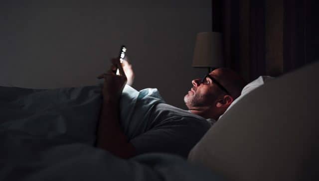 Hombre adulto tumbado en la cama, usando el móvil a oscuras con la luz del dispositivo iluminando su rostro.