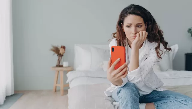 Mujer joven con expresión triste mirando el móvil mientras está sentada en una cama.