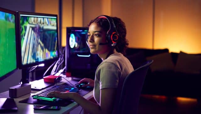Joven jugando con auriculares en un entorno gamer con varios monitores, iluminado por luces LED.