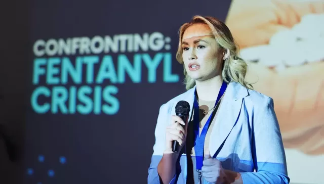Mujer hablando con micrófono frente a una pantalla donde se lee "Fentanyl Crisis", alertando sobre la emergencia sanitaria del fentanilo.