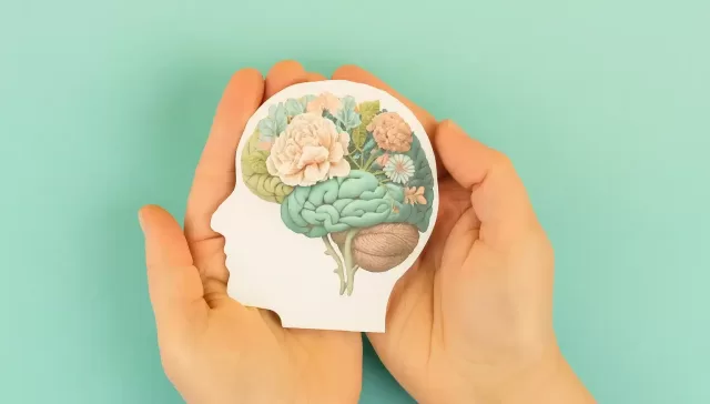 Manos sosteniendo una silueta humana con un cerebro formado por flores, símbolo del cuidado de la salud mental.